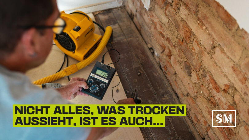 Handwerker führt in einem Rohbau mit einem digitalen Messgerät eine Feuchtigkeitsmessung am Boden durch, im Hintergrund ein gelber Bautrockner und freiliegendes Mauerwerk.